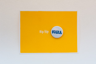 een geel verjaardagskaartje met een pin waar HOERA op staat in het blauw